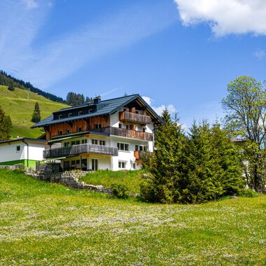 A picturesque house amidst a green meadow and surrounded by trees. In the background, gentle hills are visible under a clear blue sky.