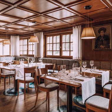 A cozy restaurant with wooden walls and elegant tablecloths. The tables are set, and there are large windows that let in plenty of light.