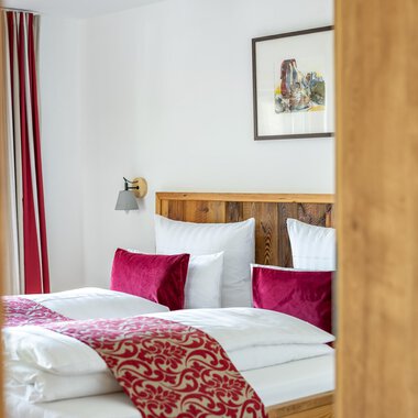 A cozy bedroom with a large bed and red pillows. The walls are light, and there is a piece of artwork in the background.