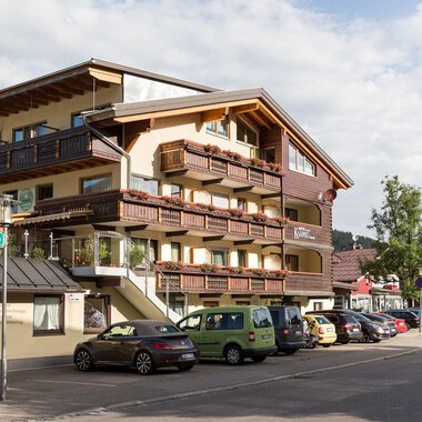 A modern building with several balconies is located on a street. Surrounded by trees and parking spaces, it exudes an inviting atmosphere.
