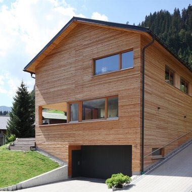 A modern wooden house in a green environment with mountains in the background. The architecture is simple and harmonizes well with nature.