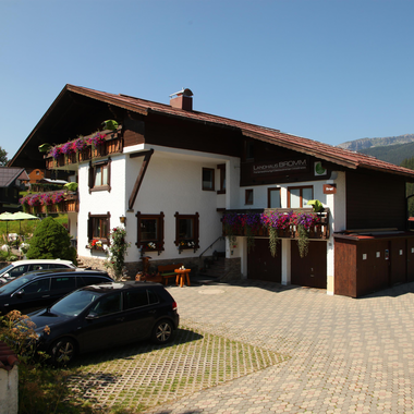 A charming building in Bavarian style with colorful flowers at the windows. The parking lot in front of the house is well-lit and has space for several cars.