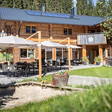A modern courtyard surrounded by green nature. On the terrace, there are tables and chairs with sun umbrellas for relaxing hours outdoors.