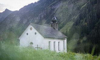 Kleine weiße Kirche mit dunklem Glockenturm vor grünen Bergen und Himmel im Hintergrund. | © Kleinwalsertal Tourismus eGen 