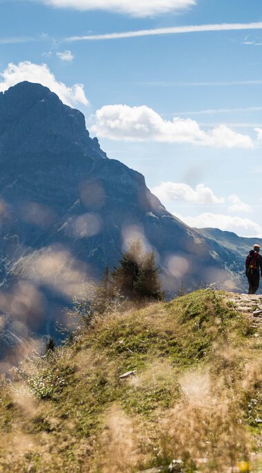 Ein Wanderer geht auf einem Pfad in den Bergen im Schwarzwald bei schönem Wetter, umgeben von grünem Gras und Bergen im Hintergrund. | © Kleinwalsertal Tourismus eGen • Fotograf: Andre Tappe