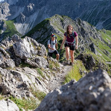 Two hikers on a narrow mountain trail with rocks and green slopes during a hike in the Alps. | © Kleinwalsertal Tourismus eGen • Photographer: Bastian Morell