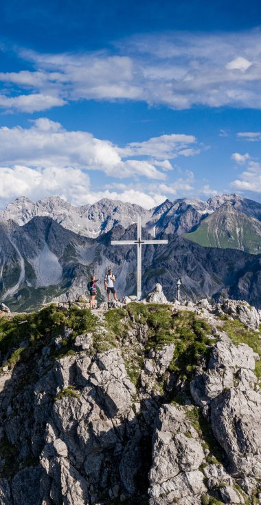 Mountain summit with cross and hikers, above the Alps, on a clear day with blue sky and clouds. | © Kleinwalsertal Tourismus eGen • Photographer: Bastian Morell