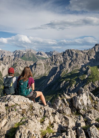 Two hikers with backpacks sitting on rocks and enjoying the mountain view of Kanzelwand summit under partly cloudy sky. | © Kleinwalsertal Tourismus eGen • Photographer: Bastian Morell