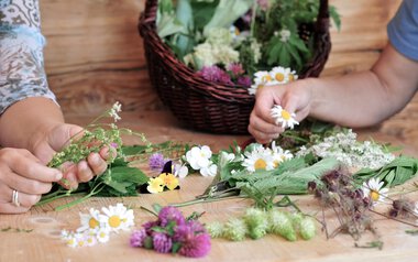 People pick herbs and flowers for a floral or herbal photoshoot at a wooden table, with a basket of flowers in the background. | © Kleinwalsertal Tourismus eGen • Photographer: Christoffer Leitner
