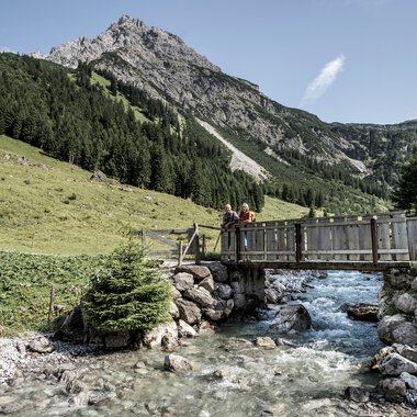 Two people on a wooden bridge over a creek in an alpine landscape with mountains in the background. | © Kleinwalsertal Tourismus eGen • Photographer: Dominik Berchtold