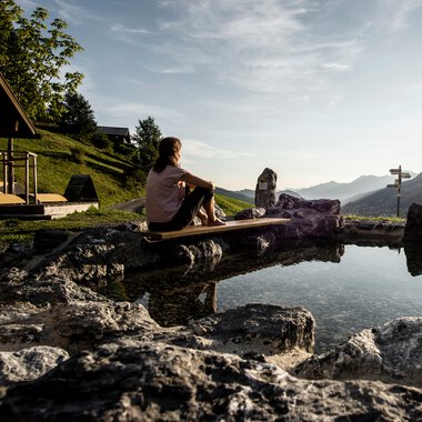 Woman sitting at the Naturerlebnisplatz Rohrweg, surrounded by rocks, overlooking mountains and sunset. | © Kleinwalsertal Tourismus eGen • Photographer: Dominik Berchtold