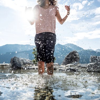 Kind läuft barfuß im Wasser an einem Naturerlebnisplatz mit Bergen im Hintergrund, Wasser spritzt beim Laufen. | © Kleinwalsertal Tourismus eGen • Fotograf: Dominik Berchtold