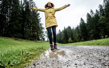 Woman in a yellow raincoat and dark pants standing on a wet forest path with arms spread out in bad weather in the forest. | © Kleinwalsertal Tourismus eGen • Photographer: Dominik Berchtold