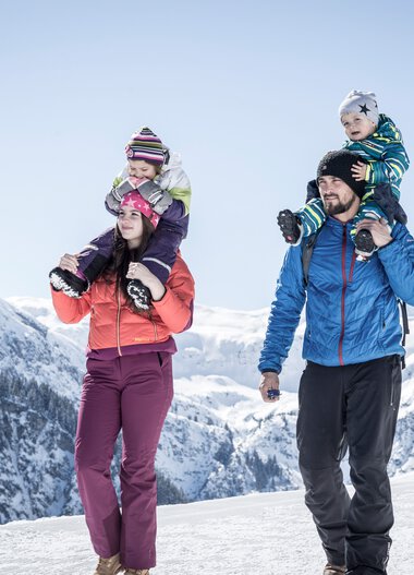 Familie beim Winterwandern in den Bergen, zwei Kinder auf den Schultern der Eltern, verschneite Gipfel im Hintergrund. | © Kleinwalsertal Tourismus eGen • Fotograf: Dominik Berchtold