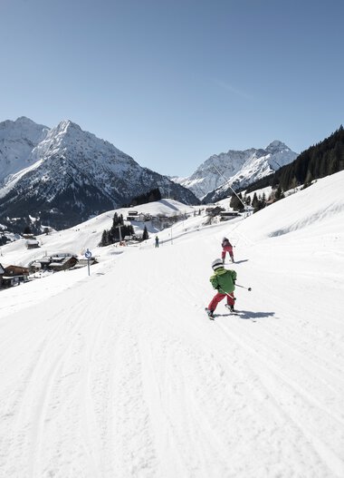 Family skiing on a snowy slope in the mountains, surrounded by winter landscape and mountain cabins. | © Kleinwalsertal Tourismus eGen • Photographer: Dominik Berchtold
