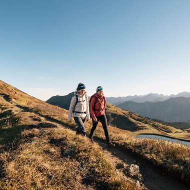 Two hikers walking on a trail in the mountains at sunset, with a mountain landscape and a lake in the background. | © Kleinwalsertal Tourismus eGen • Photographer: Hannah Bichay