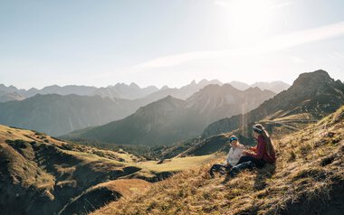 Zwei Personen sitzen entspannt auf einer Bergwiese mit Blick auf eine beeindruckende Bergkette bei Sonnenuntergang.