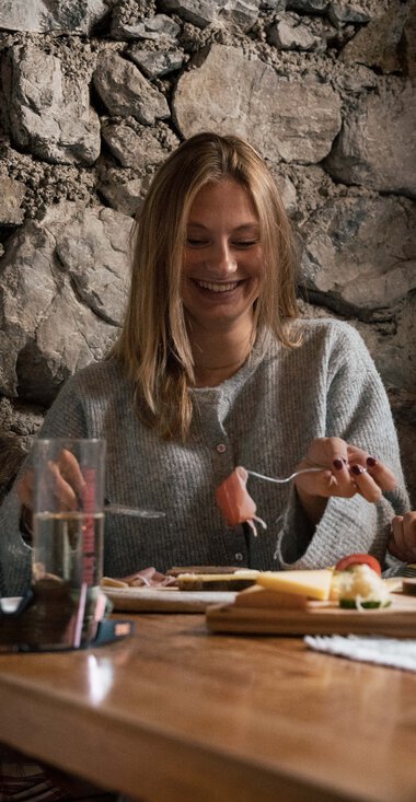 Two people are happily dining in a cozy wooden and stone cabin, laughing and engaging in conversation. | © Kleinwalsertal Tourismus eGen • Photographer: Hannah Bichay