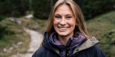 Junge Frau mit langem, blondem Haar, lachend und in einer Outdoor-Jacke, vor einem Wald- und Bach-Hintergrund, bei einem Fotoshooting in der Natur. | © Kleinwalsertal Tourismus eGen • Fotograf: Hannah Bichay