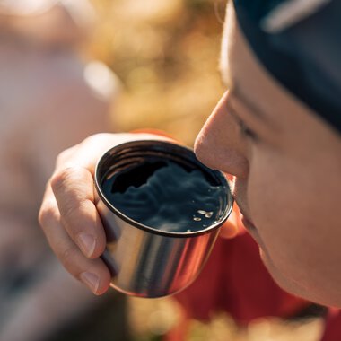 Nahaufnahme einer Person, die in der Natur ein Becher mit heißem Getränk hält und daraus trinkt, bei Sonnenlicht im Freien. | © Kleinwalsertal Tourismus eGen • Fotograf: Hannah Bichay