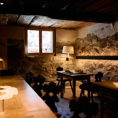 Cozy room with stone walls and ceiling, table lamps, and a chessboard on a table. | © Kleinwalsertal Tourismus eGen • Photographer: Oliver Farys