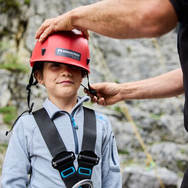 Ein Junge trägt einen Kletterhelm und wird von einem Erwachsenen für eine Bergklettertour vorbereitet. | © Kleinwalsertal Tourismus eGen • Fotograf: Oliver Farys