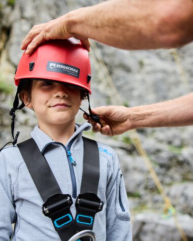 A boy wearing a climbing helmet is being prepared for a mountain climbing activity by an adult. | © Kleinwalsertal Tourismus eGen • Photographer: Oliver Farys