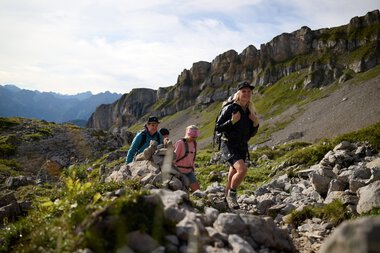 Vier Wanderer klettern auf einem felsigen Bergpfad im Gottesacker im Kleinwalsertal, mit Berggipfeln im Hintergrund an einem sonnigen Tag. | © Kleinwalsertal Tourismus eGen • Fotograf: Oliver Farys