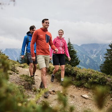 Four hikers walk along a narrow mountain trail in Kleinwalsertal with mountains and forests in the background. | © Kleinwalsertal Tourismus eGen • Photographer: Oliver Farys
