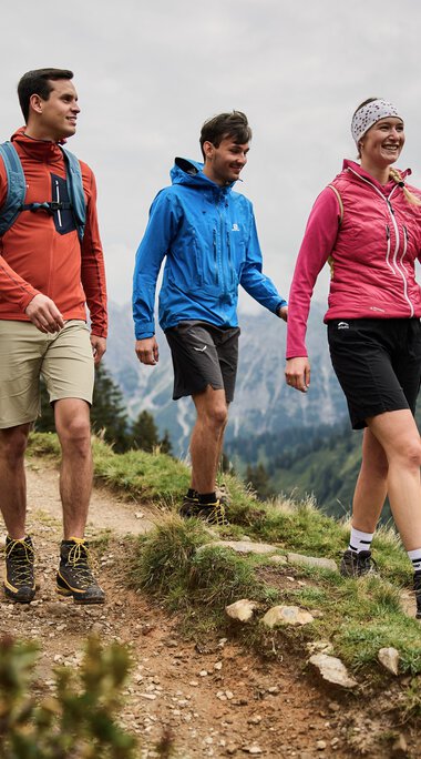 Three people hiking on a mountain trail in Kleinwalsertal, surrounded by mountains and fir trees, on a cloudy day. | © Kleinwalsertal Tourismus eGen • Photographer: Oliver Farys