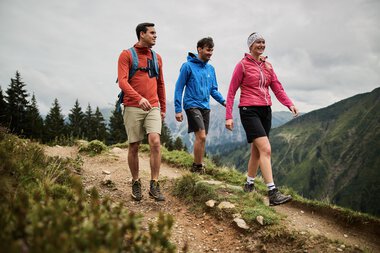 Drei Menschen wandern entlang eines Bergpfads im Kleinwalsertal, umgeben von Bergen und Tannen, bei bewölktem Himmel. | © Kleinwalsertal Tourismus eGen • Fotograf: Oliver Farys