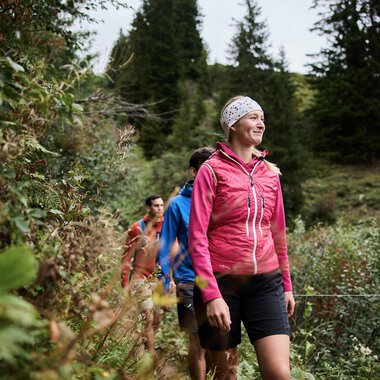 A group of hikers led by a smiling woman in a pink jacket walking through a lush, forested landscape in autumn. | © Kleinwalsertal Tourismus eGen • Photographer: Oliver Farys