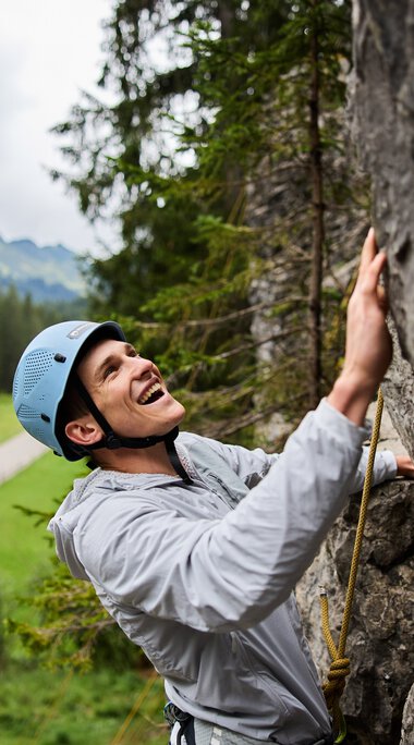 Junge Frau beim Klettern im Klettergarten, trägt Helm und Sicherheitsausrüstung, umgeben von Bergen und Wäldern. | © Kleinwalsertal Tourismus eGen • Fotograf: Oliver Farys