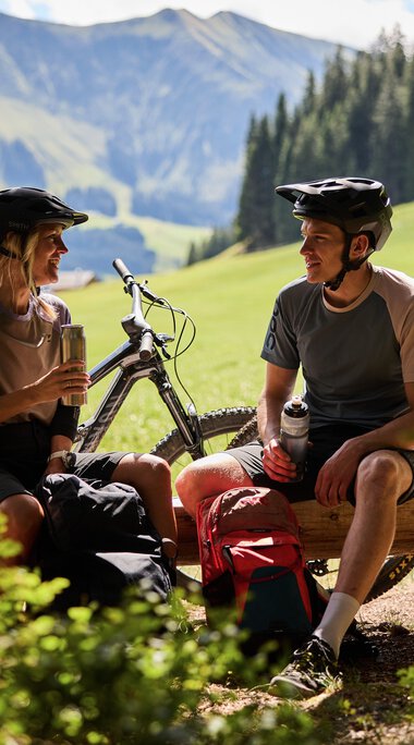 Two mountain bikers wearing helmets, sitting on a bench in a lush green area with bikes in the background during summer in Kleinwalsertal. | © Kleinwalsertal Tourismus eGen • Photographer: Oliver Farys