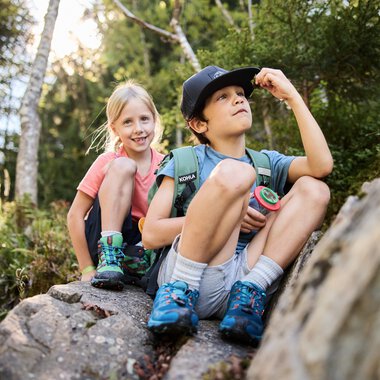Two children sitting on a rock in a forest, one looking up, the other smiling, surrounded by trees and nature. | © Kleinwalsertal Tourismus eGen • Photographer: Oliver Farys