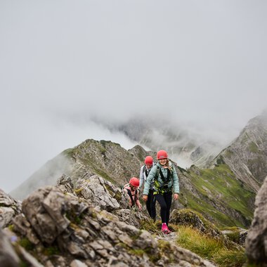 People on a via ferrata in the mountains surrounded by clouds and rocks, with Walser Mountain School logo visible. | © Kleinwalsertal Tourismus eGen • Photographer: Oliver Farys