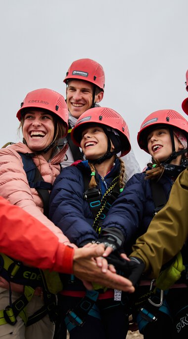 Group discussion at the via ferrata with a mountain guide and participants, all wearing helmets and safety gear, under overcast skies.