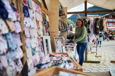 Woman selling handmade fabrics at a weekly market in Kleinwalsertal, surrounded by colorful textiles and a scenic mountain backdrop. | © Kleinwalsertal Tourismus eGen • Photographer: Oliver Farys