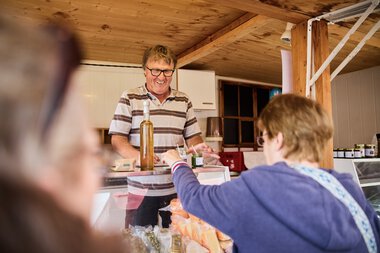 Ein Mann mit Brille und gestreiftem Hemd lächelt im Sommer 2023 am Walser Wochenmarkt im Kleinwalsertal, während eine Frau ihm ein Produkt reicht. | © Kleinwalsertal Tourismus eGen • Fotograf: Oliver Farys