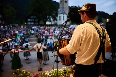 Musiker auf einer Bühne beim Sommerfest im Kleinwalsertal, im Vordergrund ein Mann mit Hut, im Hintergrund eine Tanzgruppe und eine Menschenmenge vor einer Kirche. | © Kleinwalsertal Tourismus eGen • Fotograf: Oliver Farys
