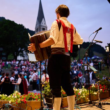 Musiker auf einer Bühne beim Alpenzauber-Festival im Kleinwalsertal, spielt Gitarre während die Menge im Hintergrund zuschaut, mit Bergkulisse und Kirche im Hintergrund. | © Kleinwalsertal Tourismus eGen • Fotograf: Oliver Farys