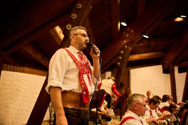 Mann in traditioneller Tracht beim Singen und Musizieren bei einem Sommerfest der Trachtenkapelle Mittelberg in einer Holzhalle. | © Kleinwalsertal Tourismus eGen • Fotograf: Oliver Farys