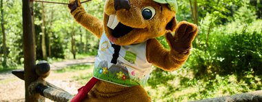 Ein faires Maskottchen in einem Wald bei einer Veranstaltung im Sommer 2024, mit grünem Hut und bunt bedruckter Weste, das auf einem Seil balanciert. | © Kleinwalsertal Tourismus eGen • Fotograf: Oliver Farys