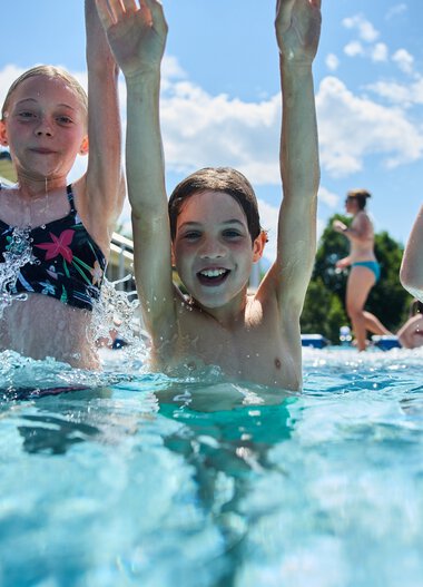 Three children enjoying themselves in the outdoor pool, laughing and playing in the water under a bright blue sky with sunlight. | © Kleinwalsertal Tourismus eGen • Photographer: Oliver Farys