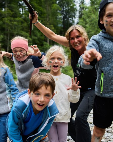 Group of children and an adult woman enjoying outdoor adventure by a river in a forest, smiling and playing. | © Kleinwalsertal Tourismus eGen • Fotograf: Oliver Farys