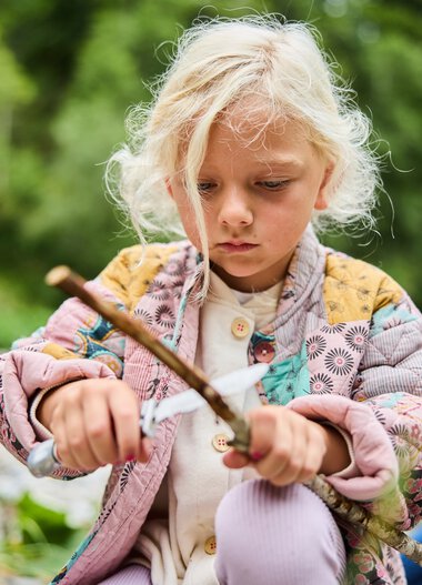 Ein junges Mädchen beschäftigt sich mit einem Stock im Grünen bei einem Outdoor-Abenteuer. | © Kleinwalsertal Tourismus eGen • Fotograf: Oliver Farys