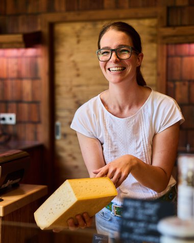 Smiling female vendor with glasses holding a piece of cheese at a farm shop in Kleinwalsertal, with wooden shelves and products in the background. | © Kleinwalsertal Tourismus eGen • Photographer: Oliver Farys