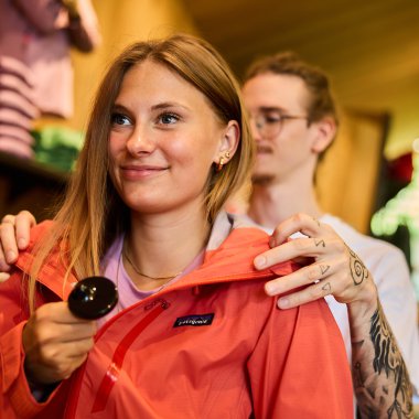 Young woman shopping in a store, wearing an orange jacket and holding a spoon in her hand. | © Kleinwalsertal Tourismus eGen • Photographer: Oliver Farys