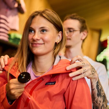 Young woman shopping in a store, wearing an orange jacket and holding a spoon in her hand. | © Kleinwalsertal Tourismus eGen • Photographer: Oliver Farys
