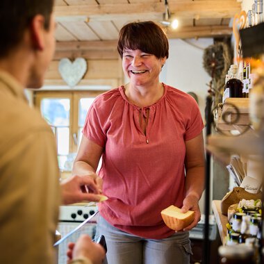 Friendly shop assistant talking to a customer, holding a slice of cheese in a store. | © Kleinwalsertal Tourismus eGen • Photographer: Oliver Farys